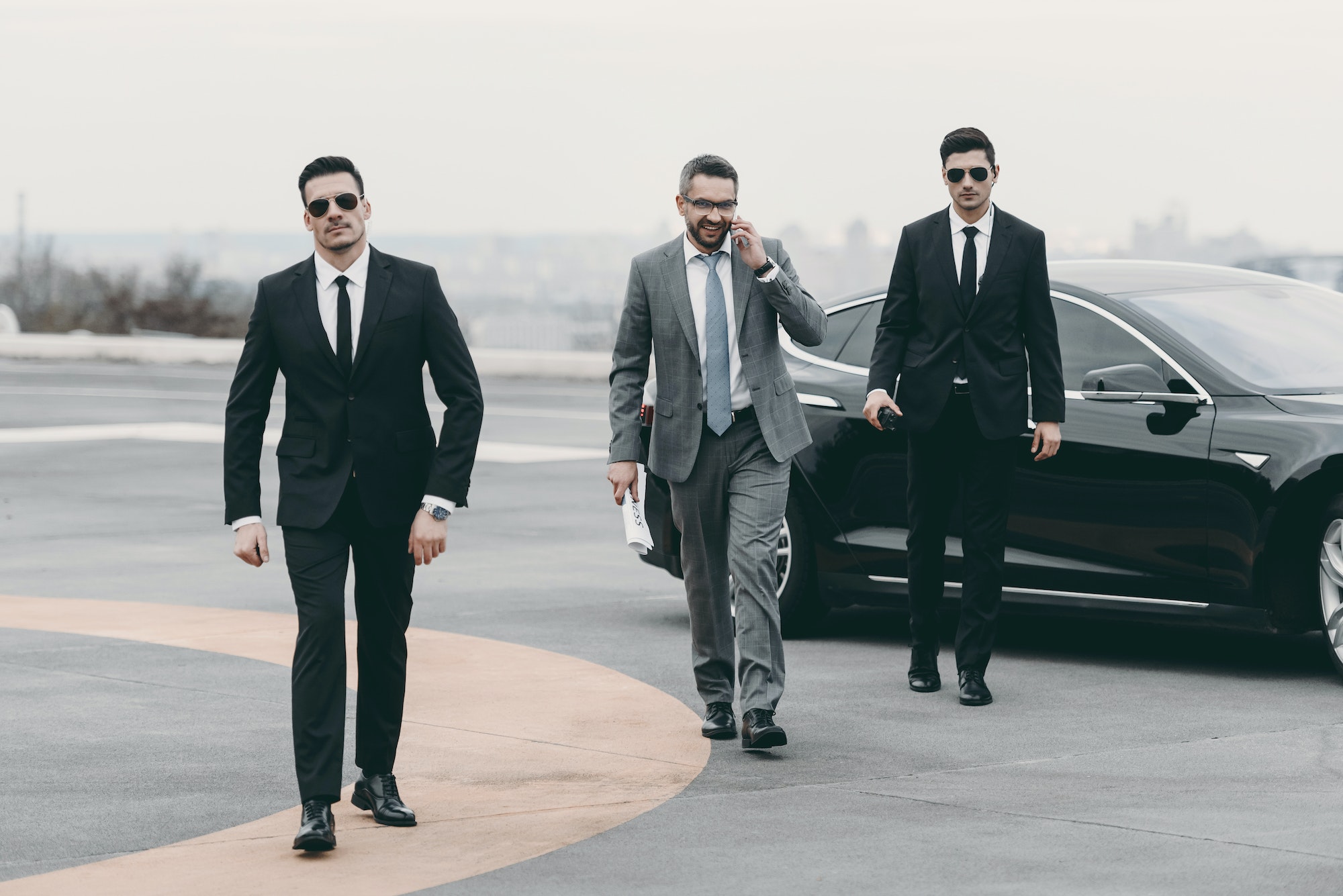 smiling businessman walking with bodyguards from car and talking by smartphone